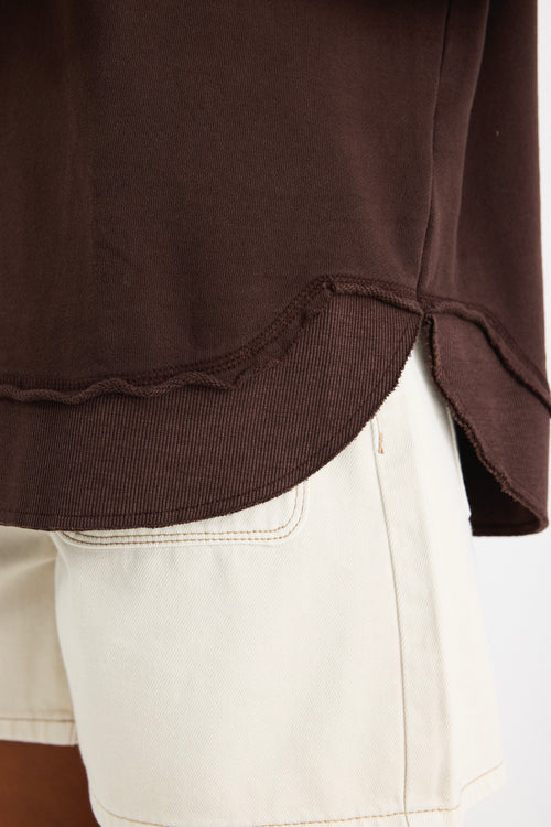 Relaxed-fit chocolate brown sweatshirt with tonal Re:Union embroidery, styled with cream shorts and gold hoop earrings.