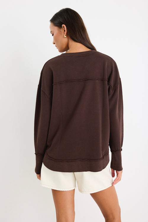 Relaxed-fit chocolate brown sweatshirt with tonal Re:Union embroidery, styled with cream shorts and gold hoop earrings.