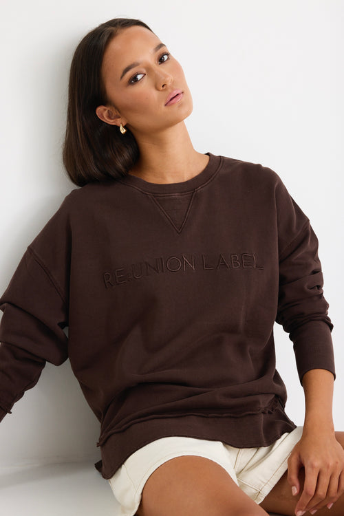 Relaxed-fit chocolate brown sweatshirt with tonal Re:Union embroidery, styled with cream shorts and gold hoop earrings.