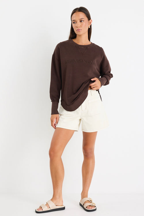 Relaxed-fit chocolate brown sweatshirt with tonal Re:Union embroidery, styled with cream shorts and gold hoop earrings.