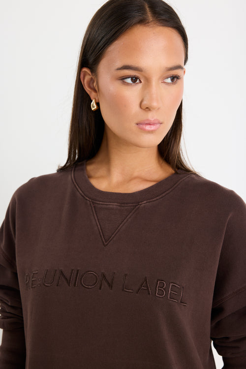 Relaxed-fit chocolate brown sweatshirt with tonal Re:Union embroidery, styled with cream shorts and gold hoop earrings.