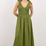 A woman modeling a sleeveless olive green midi dress with a smocked bodice and tie straps, standing against a plain white studio background.
