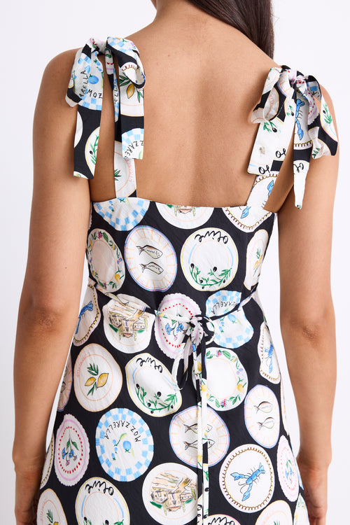 A black dress with a quirky dinner plate print, featuring strappy sleeves, a tie-back detail, and a flattering bias-cut design. Fully lined for comfort, made from a breathable viscose-nylon blend.