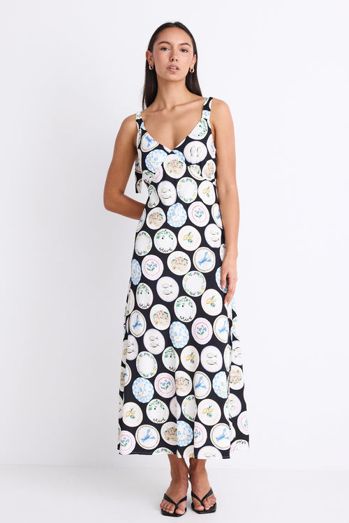 A black dress with a quirky dinner plate print, featuring strappy sleeves, a tie-back detail, and a flattering bias-cut design. Fully lined for comfort, made from a breathable viscose-nylon blend.