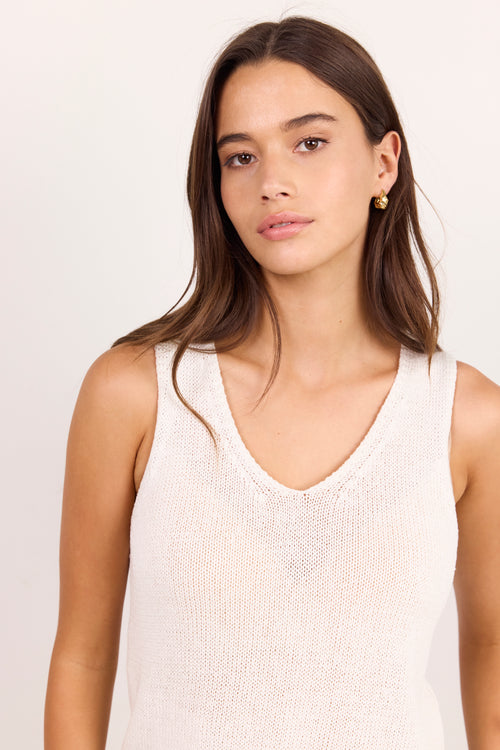 A woman with long brown hair wearing a cream-colored, textured knit sleeveless tank top, photographed against a plain light background.