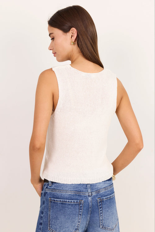 A woman shown from the back wearing a sleeveless cream knit top and blue jeans, standing against a plain off-white studio background.