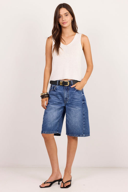 A woman modeling a pair of high-waisted, wide-leg blue denim shorts with a black belt, paired with a white sleeveless top and black sandals, against a plain white studio background.
