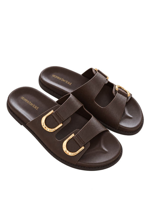 A pair of dark brown double-strap slide sandals with prominent gold-tone metal buckles, shown on a plain white background.