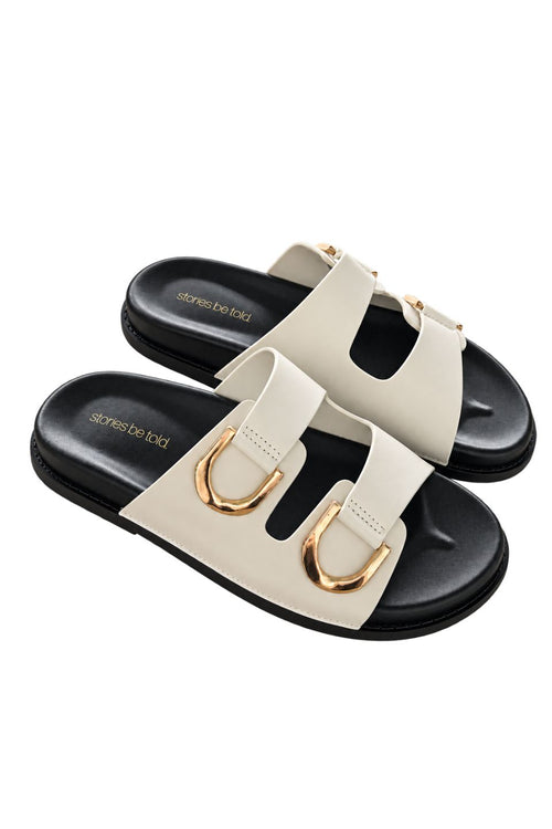 A pair of women's double-strap slide sandals by Stories Be Told, featuring off-white straps with gold-tone D-ring hardware and black soles, shown on a plain white background.