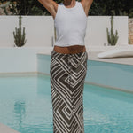 Woman wearing a khaki white print skirt and white tank in front of a pool by the sun