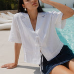 Woman wearing a whiten linen tie front top and denim mini skirt poolside in the sun