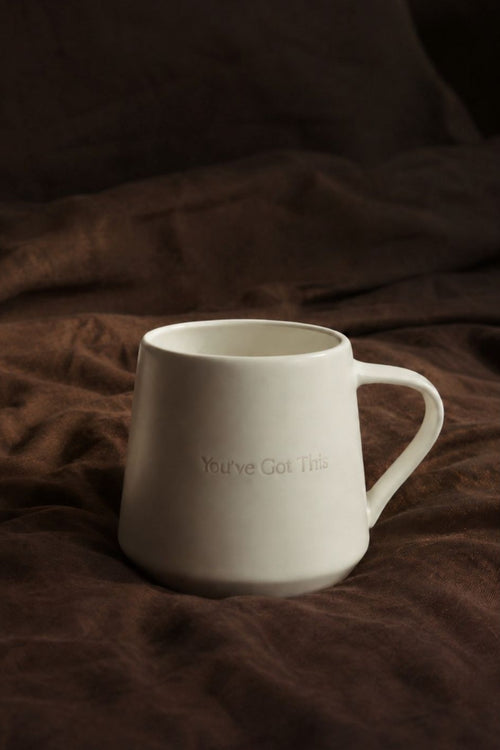A matte white ceramic mug with the phrase 'You've Got This' printed in a subtle, tonal font on the front, resting on a dark brown textured fabric background.