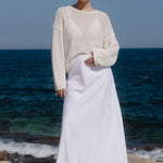Woman in a cream sweater and white skirt standing on a rocky shore with ocean in the background