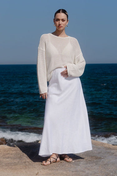 Woman in a cream sweater and white skirt standing on a rocky shore with ocean in the background