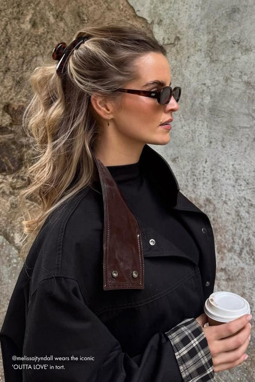 Woman wearing sunglasses and a black coat with leather accents, holding a coffee cup against a stone wall.