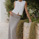 Woman in a white sleeveless top and checkered pants standing outdoors with greenery in the background