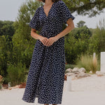 Woman in a navy blue dress with white polka dots standing by a pool with greenery in the background