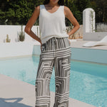 Woman wearing khaki and white print pants and a white top infront of a pool