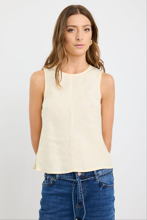 Woman wearing a sleeveless butter-yellow linen shell top with a round neckline and relaxed bias cut, styled with deep blue jeans.