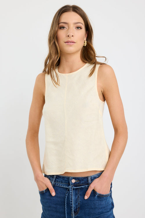 Woman wearing a sleeveless butter-yellow linen shell top with a round neckline and relaxed bias cut, styled with deep blue jeans.