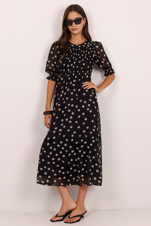 A woman modeling a black midi dress with a beige polka dot pattern, featuring short puff sleeves and a round neckline, set against a plain white studio background.