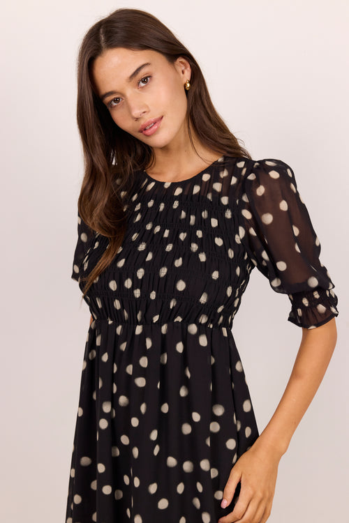 A woman wearing a black midi dress with beige polka dots, featuring a smocked bodice and sheer puff sleeves, photographed against a plain light background.