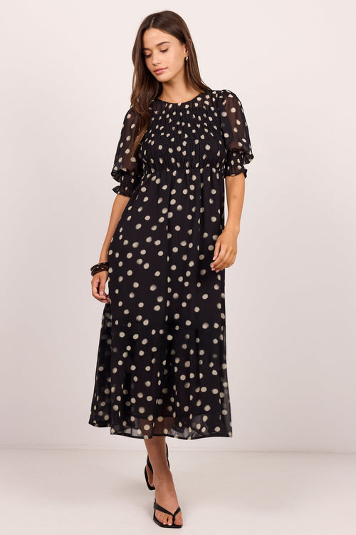 A woman modeling a black midi dress with beige polka dots, featuring puff sleeves and a tiered ruffle hem, set against a plain light background.
