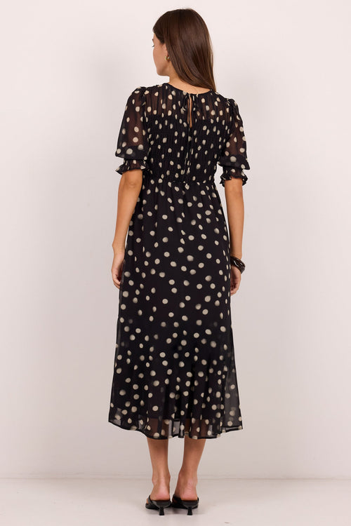 A woman viewed from the back wearing a black midi dress with a white polka dot pattern, short puff sleeves, and a back keyhole detail, paired with black heels against a plain white studio background.