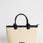 A structured tote bag in cream-colored faux leather with black handles, trim, and a small black front plaque, shown against a plain light gray background.