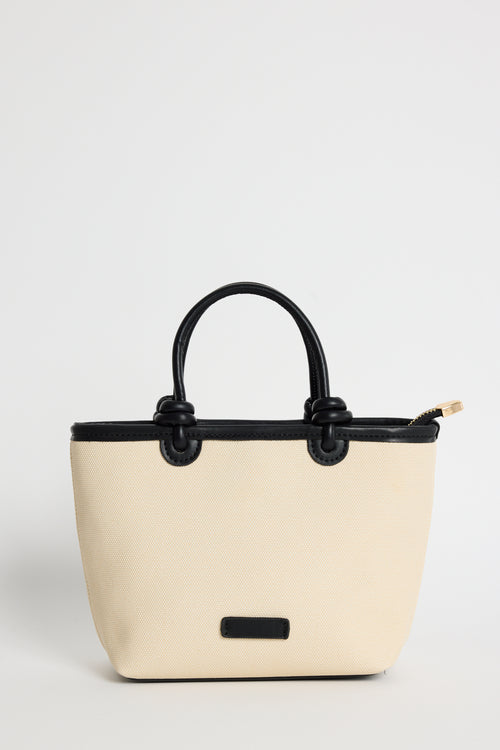 A structured tote bag in cream-colored faux leather with black handles, trim, and a small black front plaque, shown against a plain light gray background.