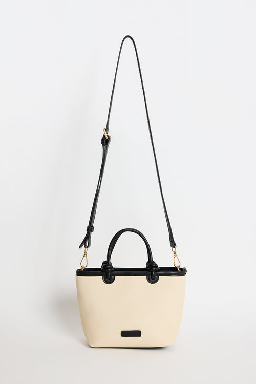 A structured cream-colored handbag with black trim, double top handles, and a long black shoulder strap, photographed against a clean white background.