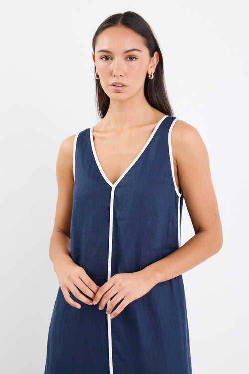 A sleeveless navy dress with ecru contrast piping down the front, featuring side pockets and a relaxed fit. Made from 100% linen for a breathable, comfortable feel.