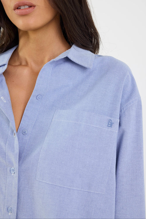 Woman wearing a light blue button-up shirt with a visible brand logo on a white background