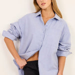 A woman modeling a light blue button-up shirt with rolled sleeves and a black maxi skirt, standing against a plain off-white studio background.