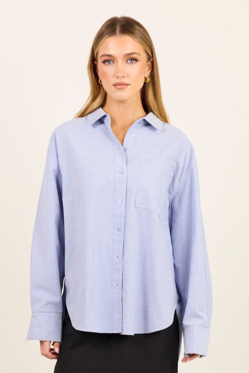 A woman modeling a light blue long-sleeve button-up shirt paired with a black maxi skirt and black sandals, standing against a plain off-white studio background.