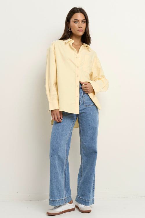 Woman wearing an oversized lemon yellow button-down shirt with wide cuffs and a curved hem, styled with light blue wide-leg jeans and white sneakers.