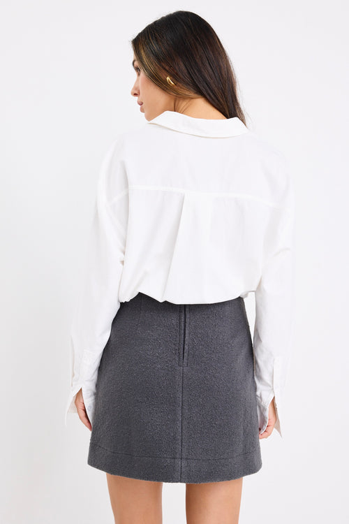 Woman wearing a dark charcoal grey mini skirt with a white oversized shirt and black sneakers, showing a clean, minimal outfit with street-style flair.