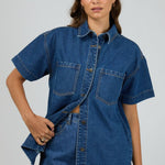 Woman wears a dark blue denim shirt with short sleeves and front patch pockets, styled with matching high-waisted denim shorts. The shirt is unbuttoned at the bottom for a relaxed, effortless look.