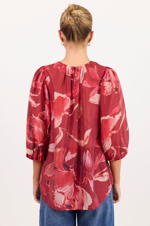 A woman viewed from behind wearing a red floral long-sleeve blouse with puff sleeves and blue jeans, standing against a plain white background.