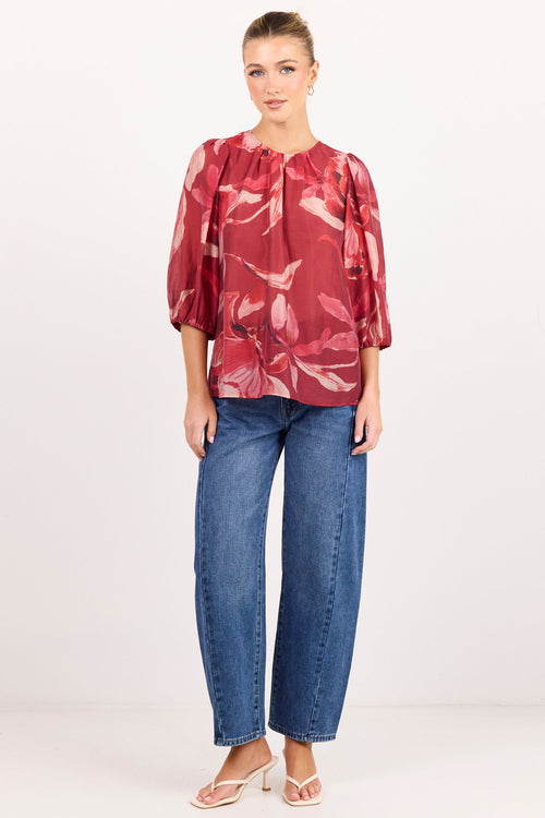 A woman modeling a red and pink abstract patterned blouse with three-quarter puffed sleeves, paired with blue jeans and light pink sandals, standing against a plain white studio background.