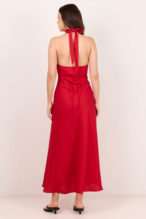 A woman viewed from the back wearing a sleeveless red halter maxi dress with a tied back neckline and a matching waist tie, paired with black mule heels against a plain light beige studio background.