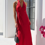 A woman wearing a vibrant red halter-neck maxi dress and black sunglasses stands on a white tiled patio in front of a white building with arched windows. She is holding a small black handbag.