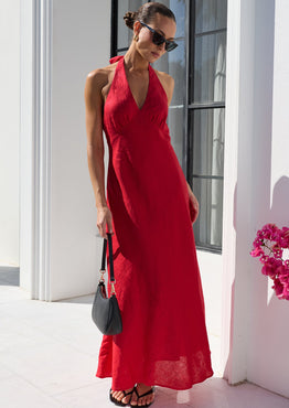 A woman wearing a vibrant red halter-neck maxi dress and black sunglasses stands on a white tiled patio in front of a white building with arched windows. She is holding a small black handbag.
