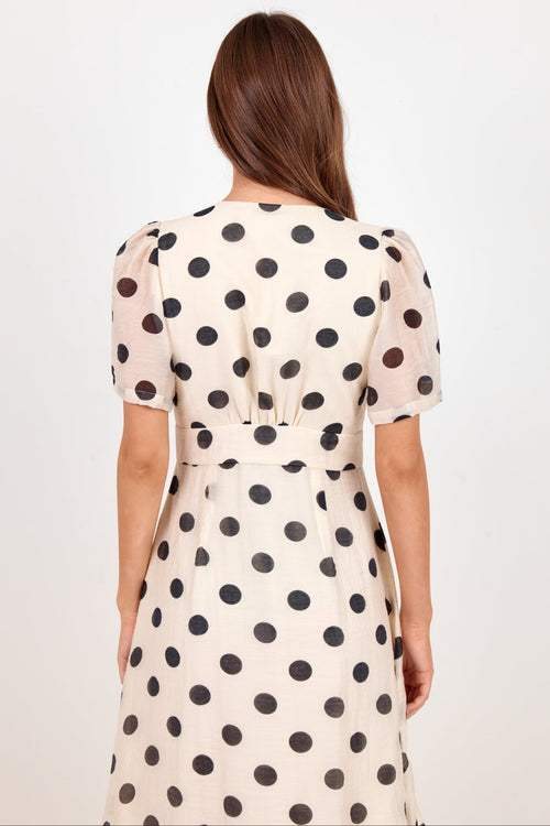 A woman viewed from the back wearing a short-sleeve cream dress with large black polka dots, set against a plain white studio background.