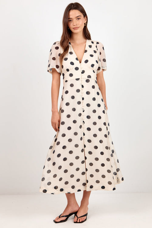 A woman modeling a cream midi dress with black polka dots, short flutter sleeves, and a V-neckline, paired with black flat sandals against a plain white studio background.
