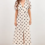 A woman modeling a cream-colored midi dress with black polka dots, featuring short puff sleeves, a V-neckline, and a front slit, set against a plain white studio background.