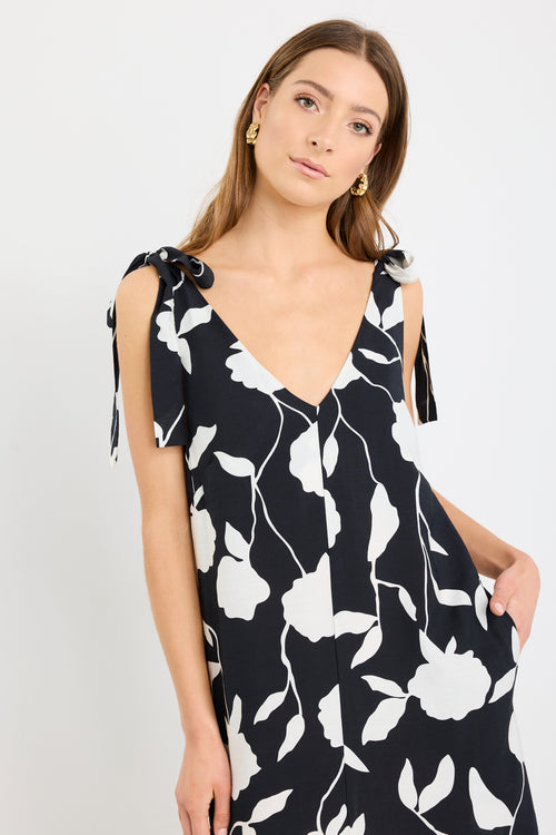 Woman wearing a sleeveless black midi dress with large white floral print, tie details at the shoulders, and a relaxed flowing fit styled with black sandals.