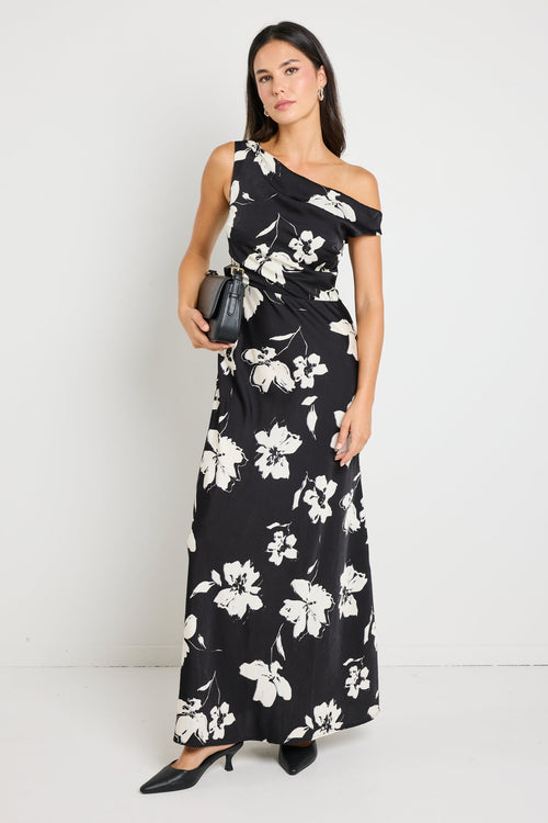 Woman wearing a one-shoulder black floral maxi dress, styled with black heels and a clutch for an elegant evening look.