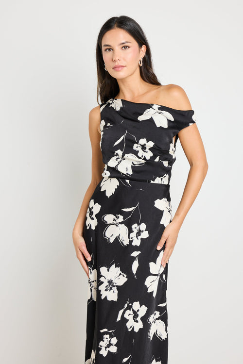 Woman wearing a one-shoulder black floral maxi dress, styled with black heels and a clutch for an elegant evening look.