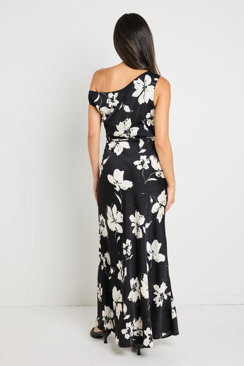 Woman wearing a one-shoulder black floral maxi dress, styled with black heels and a clutch for an elegant evening look.